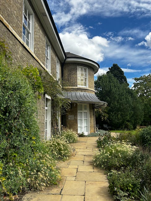 A tranquil garden scene featuring a neatly maintained lawn with lush, bright green grass bordered by a flower bed filled with leafy green plants and blooming shrubs. On the left side, there is a woode