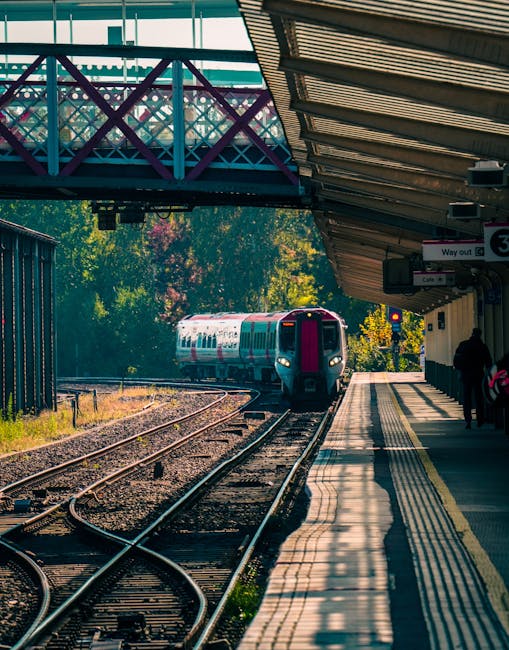 A train arriving at Cheam Station platform with passengers waiting under a metallic canopy. The platform has tactile paving along its edge and is lined with a row of signs and schedule information. Be